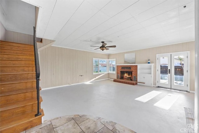 unfurnished living room featuring wooden walls, stairs, a fireplace, french doors, and a ceiling fan