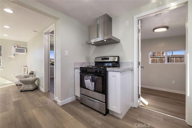 kitchen with light wood finished floors, gas range oven, a wall mounted air conditioner, white cabinetry, and wall chimney exhaust hood