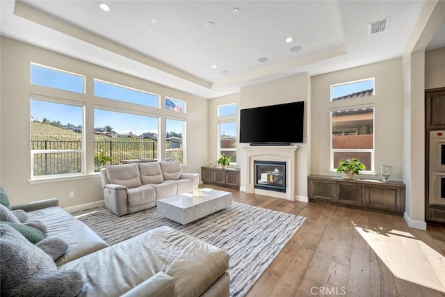 living room featuring a glass covered fireplace, light wood-style flooring, a tray ceiling, and visible vents