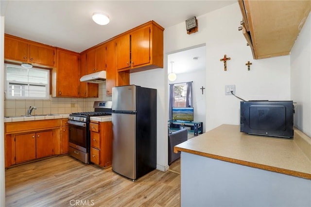 kitchen with backsplash, under cabinet range hood, brown cabinets, appliances with stainless steel finishes, and light wood-style floors