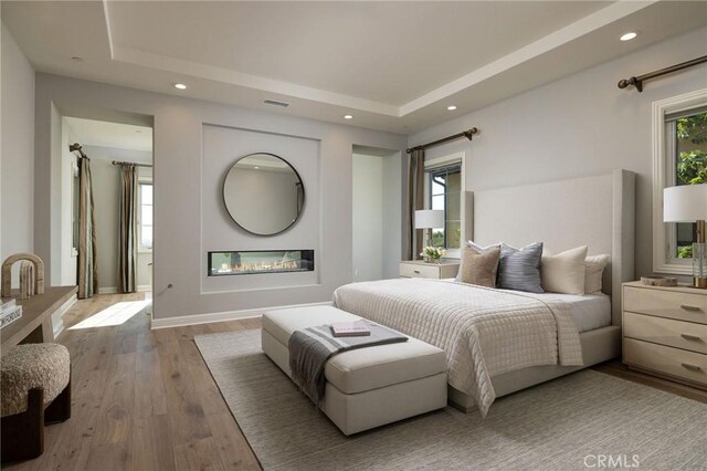 bedroom with a glass covered fireplace, a raised ceiling, recessed lighting, and wood finished floors