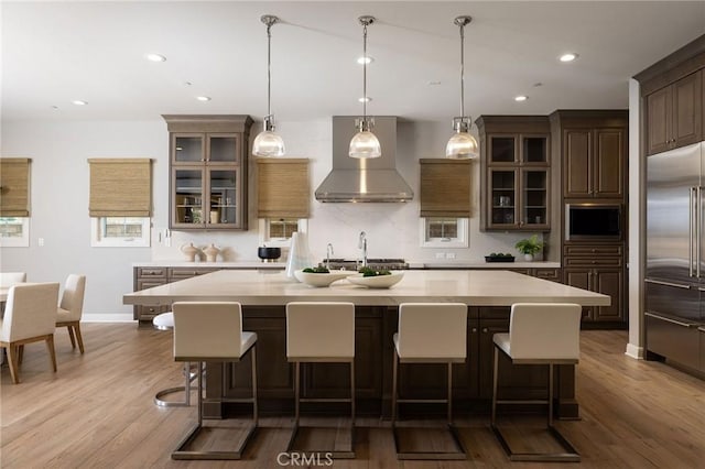 kitchen with a kitchen island with sink, wall chimney range hood, wood finished floors, light countertops, and built in appliances