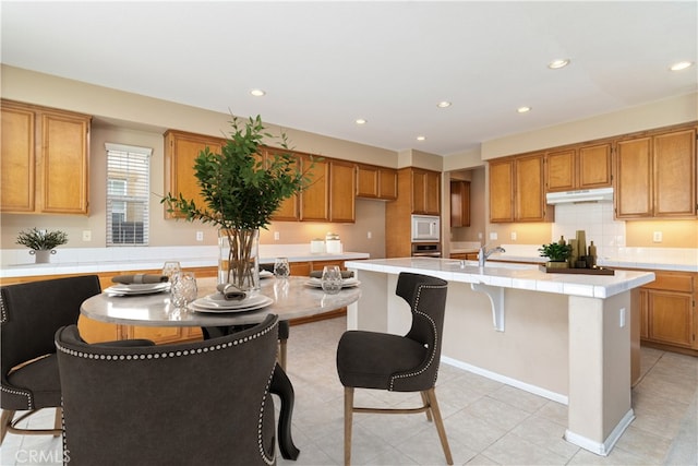 kitchen featuring brown cabinetry, an island with sink, white microwave, and under cabinet range hood