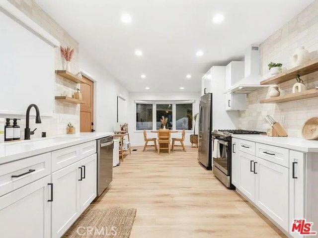 kitchen with open shelves, light wood-style flooring, a sink, appliances with stainless steel finishes, and wall chimney exhaust hood