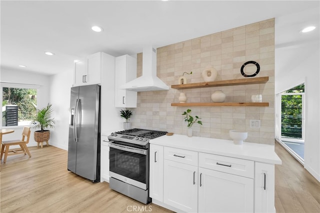 kitchen with wall chimney range hood, backsplash, light wood-style floors, and appliances with stainless steel finishes