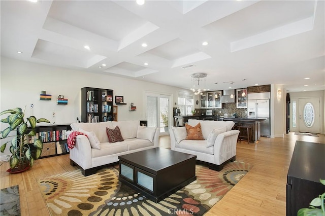 living room featuring coffered ceiling, a notable chandelier, a healthy amount of sunlight, and light wood finished floors