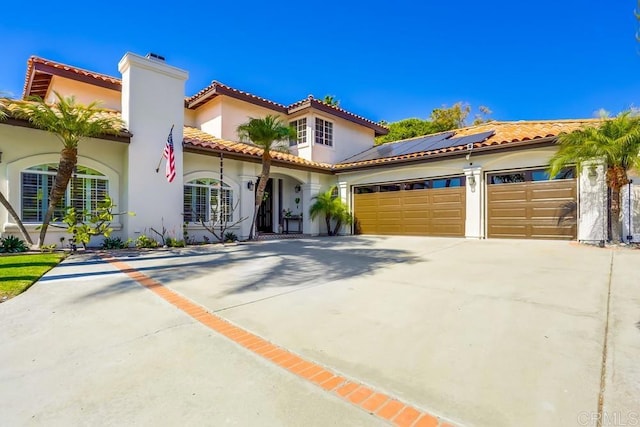 mediterranean / spanish home featuring stucco siding, driveway, roof mounted solar panels, an attached garage, and a chimney