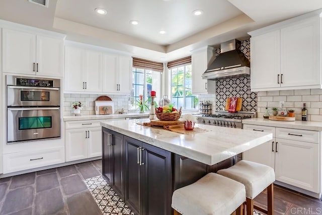 kitchen featuring stainless steel double oven, a tray ceiling, white cabinetry, and wall chimney range hood