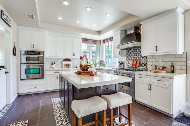 kitchen featuring a raised ceiling, wall chimney range hood, a breakfast bar area, and stainless steel appliances