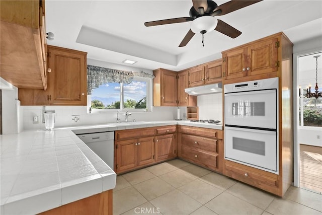 kitchen with white appliances, tile countertops, brown cabinetry, a sink, and under cabinet range hood