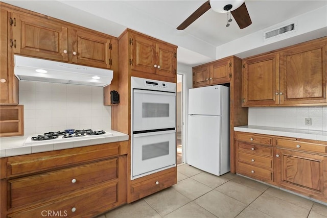 kitchen with visible vents, tile counters, under cabinet range hood, brown cabinetry, and white appliances