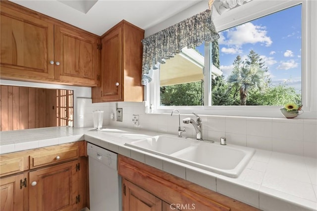 kitchen with a sink, brown cabinetry, tile counters, and white dishwasher