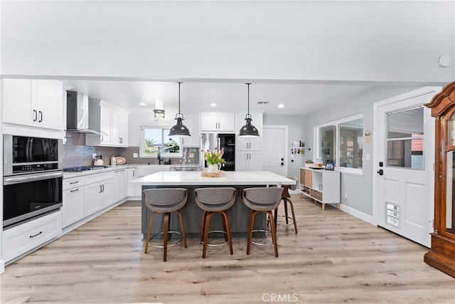 kitchen featuring light wood-style flooring, a kitchen island, white cabinetry, appliances with stainless steel finishes, and wall chimney exhaust hood