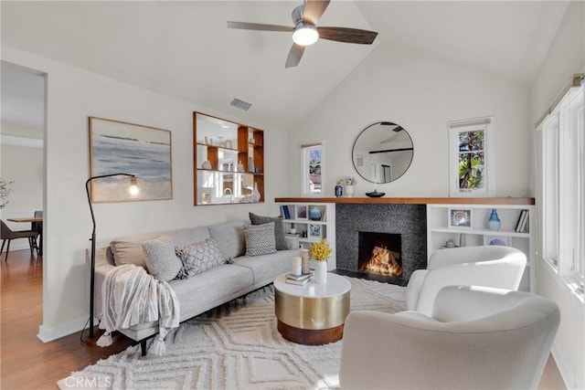 living area with visible vents, a warm lit fireplace, wood finished floors, high vaulted ceiling, and a ceiling fan