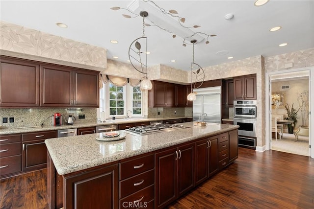 kitchen featuring wallpapered walls, light stone countertops, dark wood finished floors, an island with sink, and stainless steel appliances