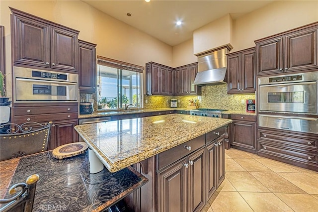 kitchen with light tile patterned floors, decorative backsplash, appliances with stainless steel finishes, a warming drawer, and wall chimney exhaust hood