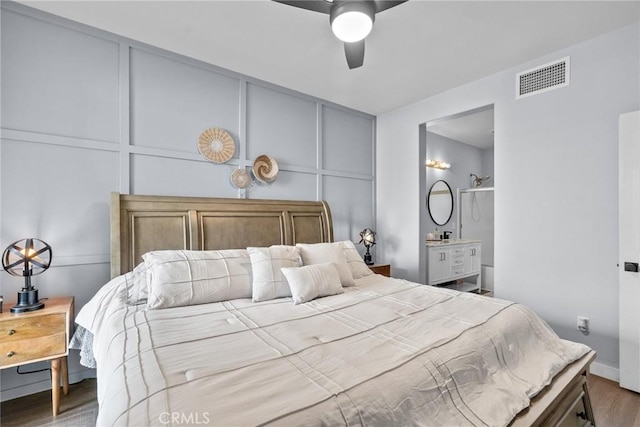 bedroom featuring wood finished floors, visible vents, ensuite bath, ceiling fan, and a decorative wall