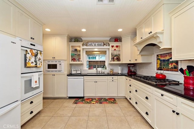 kitchen featuring dark countertops, light tile patterned floors, decorative backsplash, white appliances, and a sink