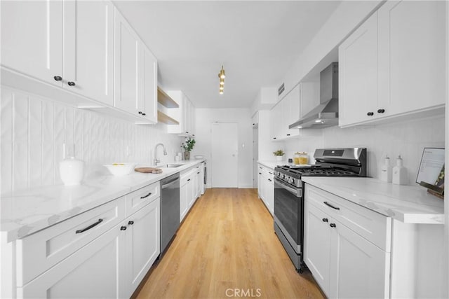 kitchen with wall chimney range hood, light wood-style flooring, stainless steel appliances, white cabinetry, and a sink