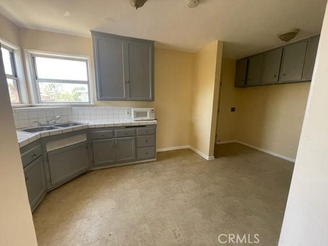 kitchen featuring baseboards, tile counters, decorative backsplash, gray cabinets, and a sink