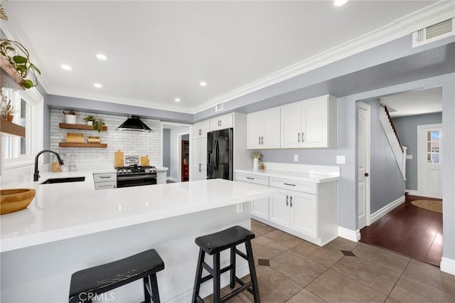 kitchen with a sink, black fridge with ice dispenser, stainless steel gas range oven, crown molding, and wall chimney range hood