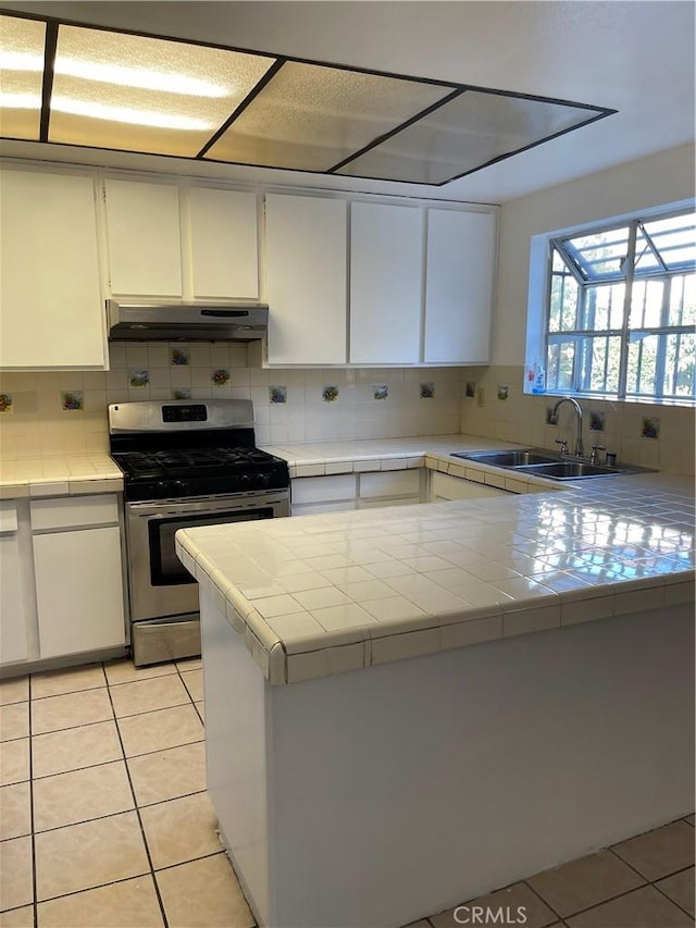 kitchen featuring under cabinet range hood, a sink, tile countertops, stainless steel range with gas cooktop, and light tile patterned flooring