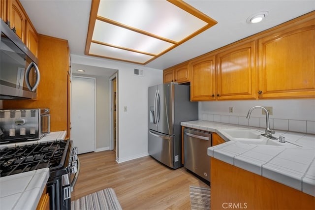 kitchen featuring light wood-style flooring, a sink, stainless steel appliances, tile counters, and brown cabinets