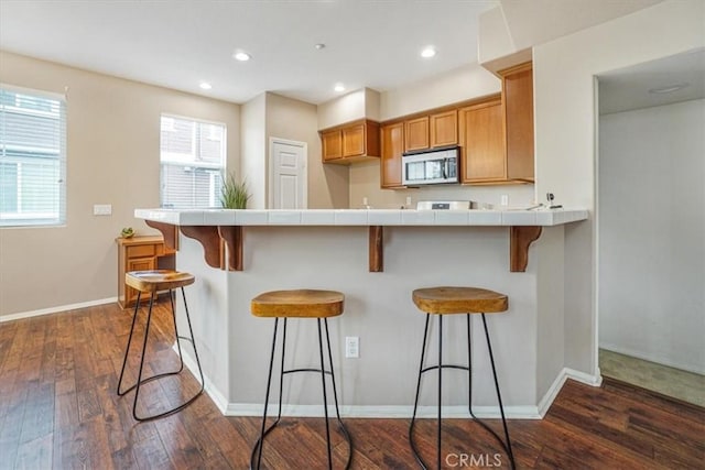 kitchen with tile countertops, dark wood finished floors, a kitchen bar, stainless steel microwave, and brown cabinets