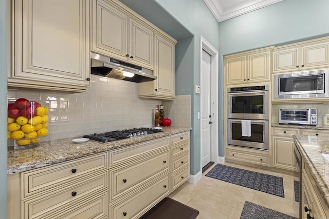 kitchen with tasteful backsplash, under cabinet range hood, ornamental molding, cream cabinetry, and stainless steel appliances