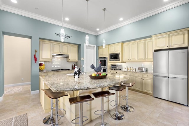 kitchen with under cabinet range hood, cream cabinets, stainless steel appliances, and light stone countertops