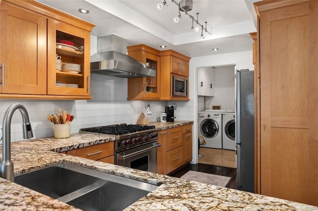 kitchen with brown cabinetry, a tray ceiling, appliances with stainless steel finishes, exhaust hood, and washing machine and dryer