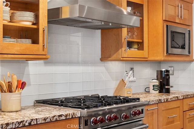 kitchen with decorative backsplash, light stone counters, appliances with stainless steel finishes, wall chimney exhaust hood, and glass insert cabinets