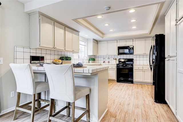 kitchen with a peninsula, black appliances, tile counters, a raised ceiling, and light wood-type flooring