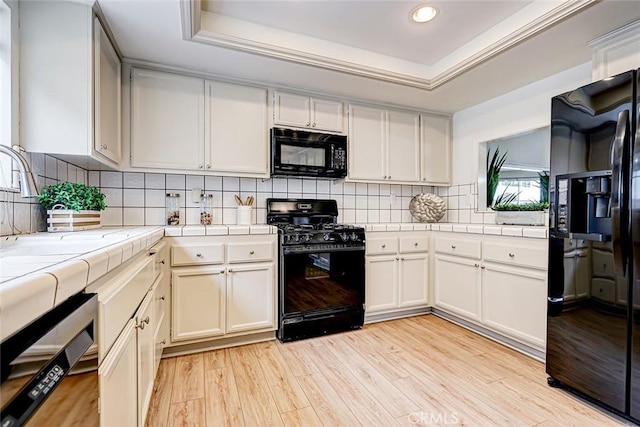 kitchen with black appliances, tile countertops, light wood-style floors, and a tray ceiling