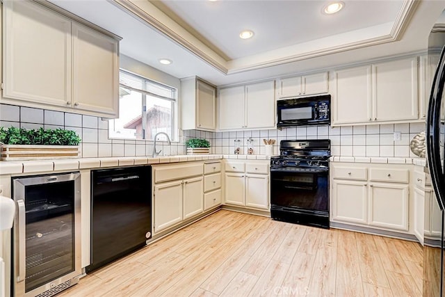 kitchen with light wood-type flooring, black appliances, beverage cooler, a tray ceiling, and tile countertops