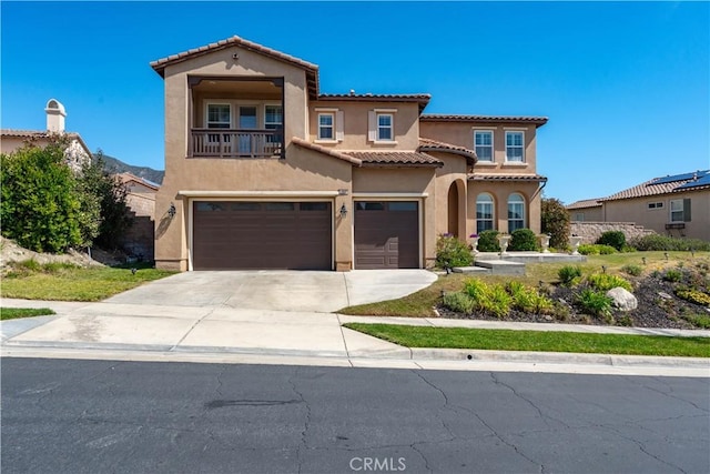 mediterranean / spanish house with a balcony, driveway, stucco siding, a garage, and a tiled roof