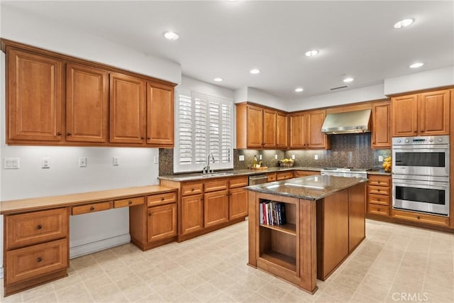 kitchen with wall chimney range hood, brown cabinetry, light floors, and stainless steel appliances