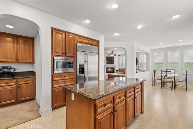 kitchen with dark stone countertops, built in appliances, a warm lit fireplace, and brown cabinetry