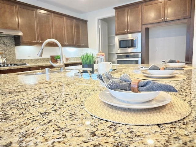 kitchen with a sink, under cabinet range hood, stainless steel appliances, decorative backsplash, and light stone countertops