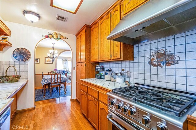 kitchen featuring stainless steel gas range oven, under cabinet range hood, tile countertops, brown cabinetry, and light wood finished floors