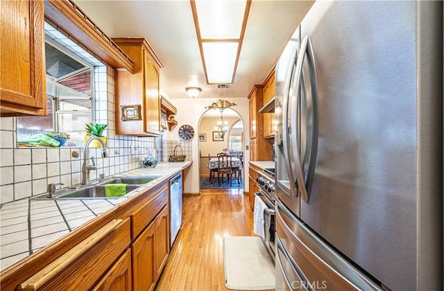 kitchen featuring tile countertops, brown cabinetry, arched walkways, a sink, and appliances with stainless steel finishes