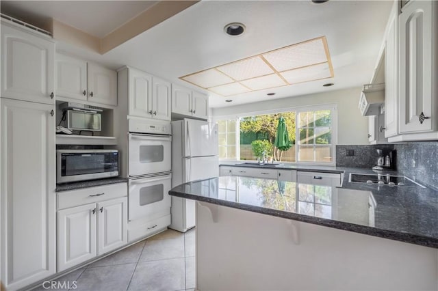 kitchen with white appliances, dark stone countertops, light tile patterned floors, a sink, and white cabinetry