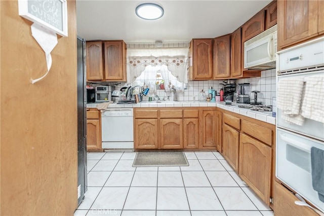 kitchen with a sink, backsplash, white appliances, brown cabinetry, and light tile patterned floors