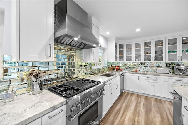 kitchen featuring light wood finished floors, a sink, stainless steel appliances, white cabinetry, and wall chimney range hood