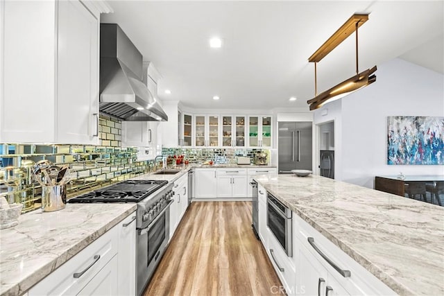 kitchen with tasteful backsplash, glass insert cabinets, wall chimney range hood, built in appliances, and light wood-type flooring