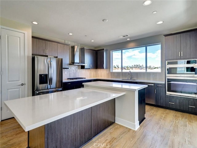 kitchen with a sink, stainless steel appliances, light countertops, a large island, and wall chimney range hood