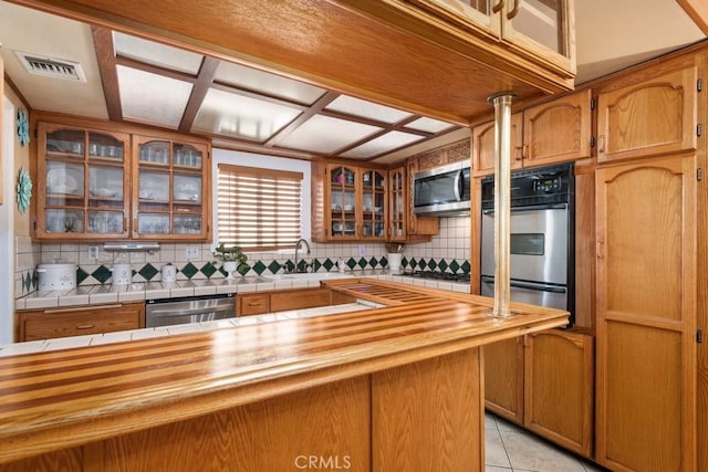 kitchen featuring visible vents, backsplash, appliances with stainless steel finishes, light tile patterned floors, and glass insert cabinets