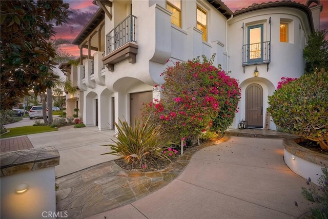 exterior entry at dusk with stucco siding, a tiled roof, concrete driveway, and an attached garage