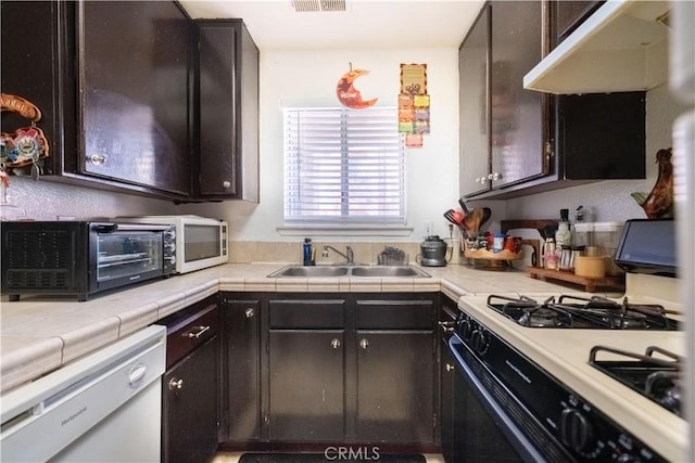 kitchen with under cabinet range hood, tile countertops, gas range oven, white dishwasher, and a sink