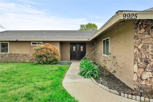 property entrance featuring stucco siding, stone siding, a lawn, and a shingled roof